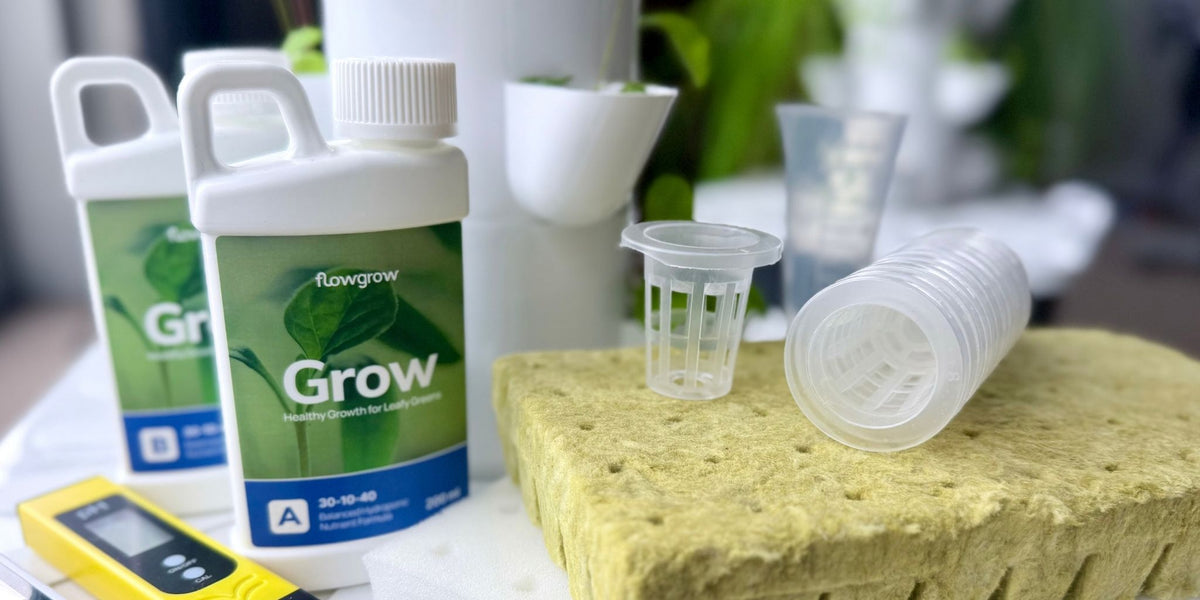 Two FlowGrow Grow nutrient bottles with white caps and green labels, alongside net pots and rockwool planting sponge, arranged on a FlowGrow Tower with a blurred indoor background.